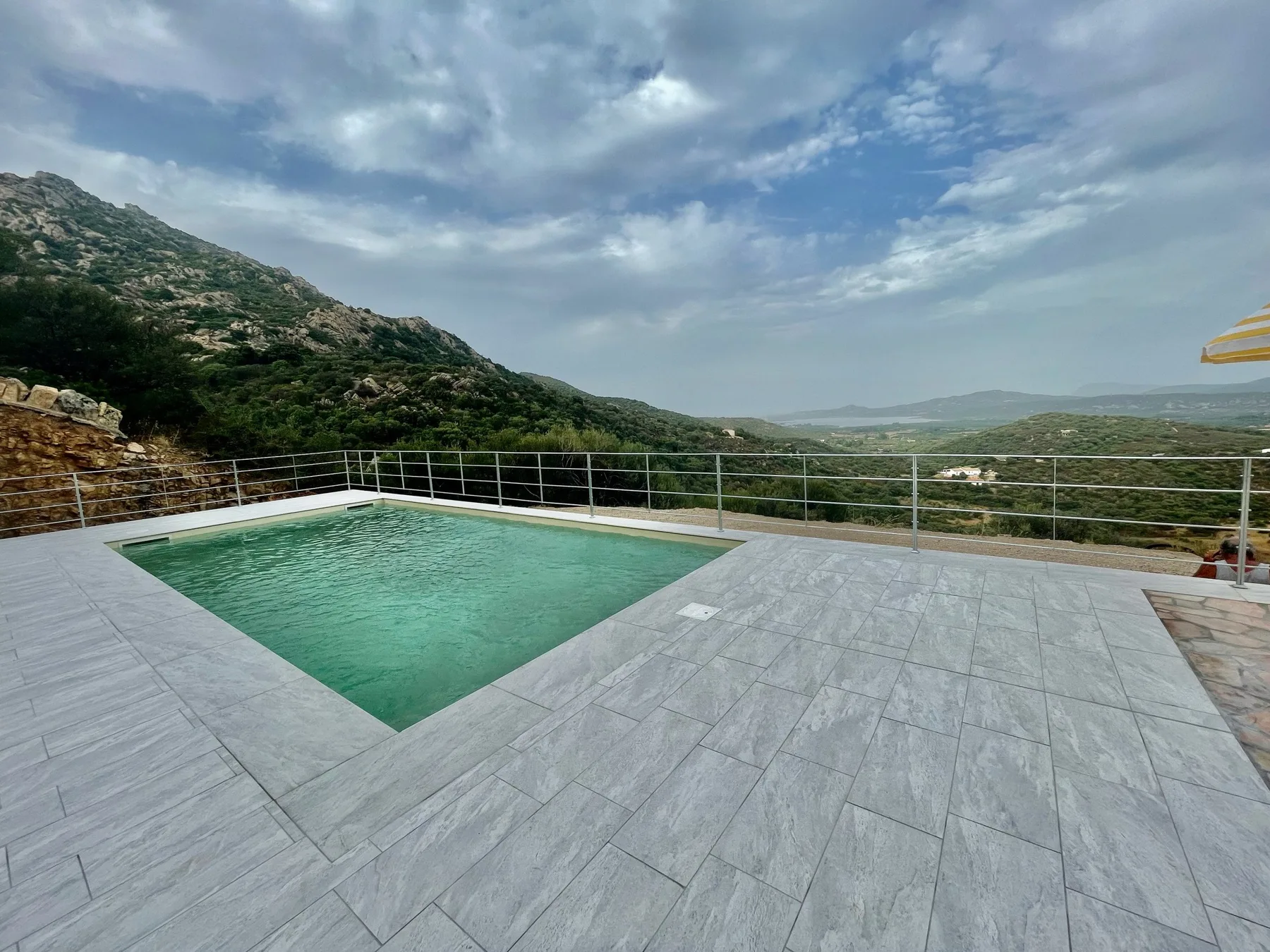 An outdoor swimming pool overlooks a green mountain valley beneath a cloudy sky.