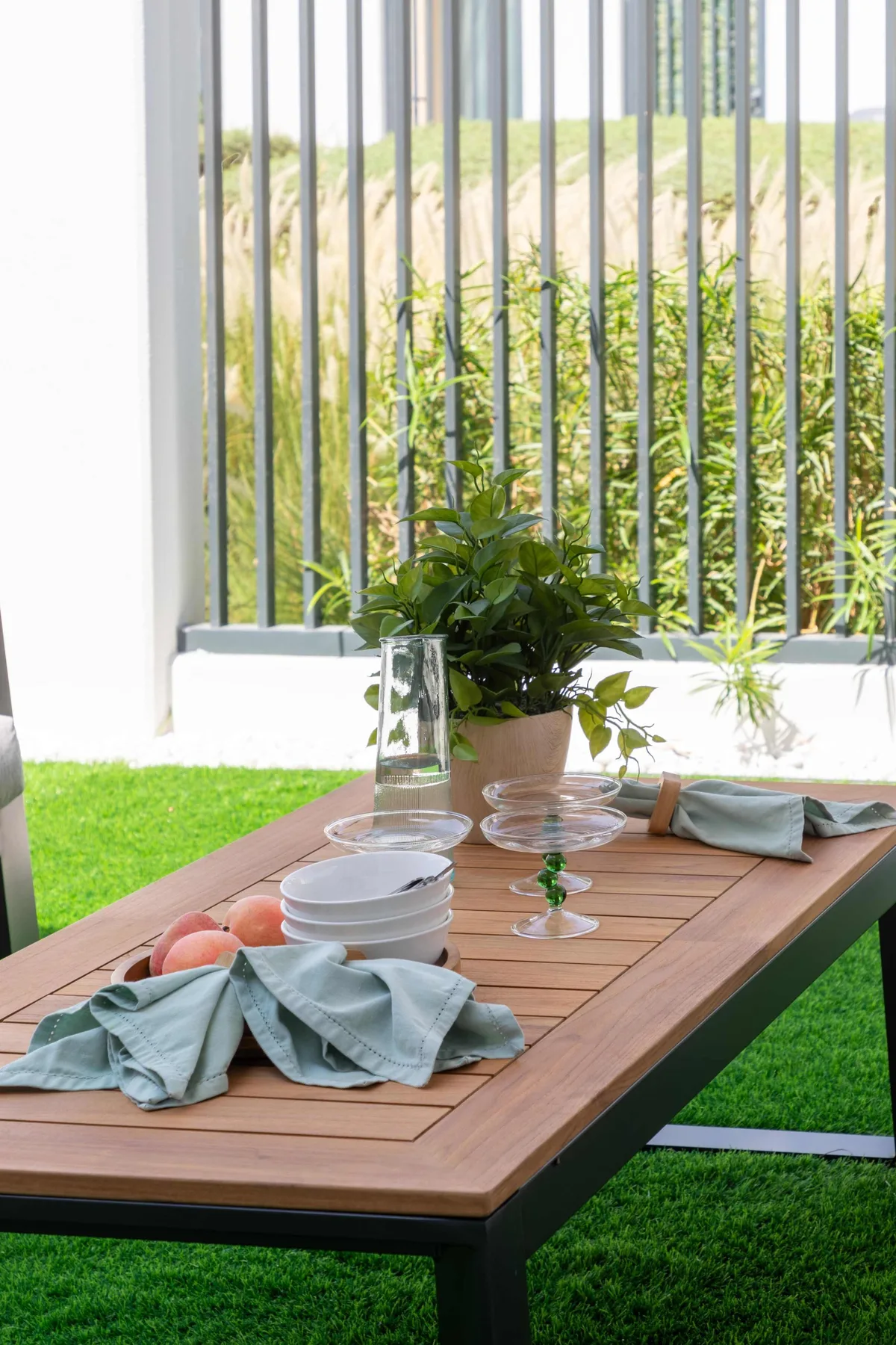 An outdoor dining table is set with dishes and a potted plant beside a grassy area.