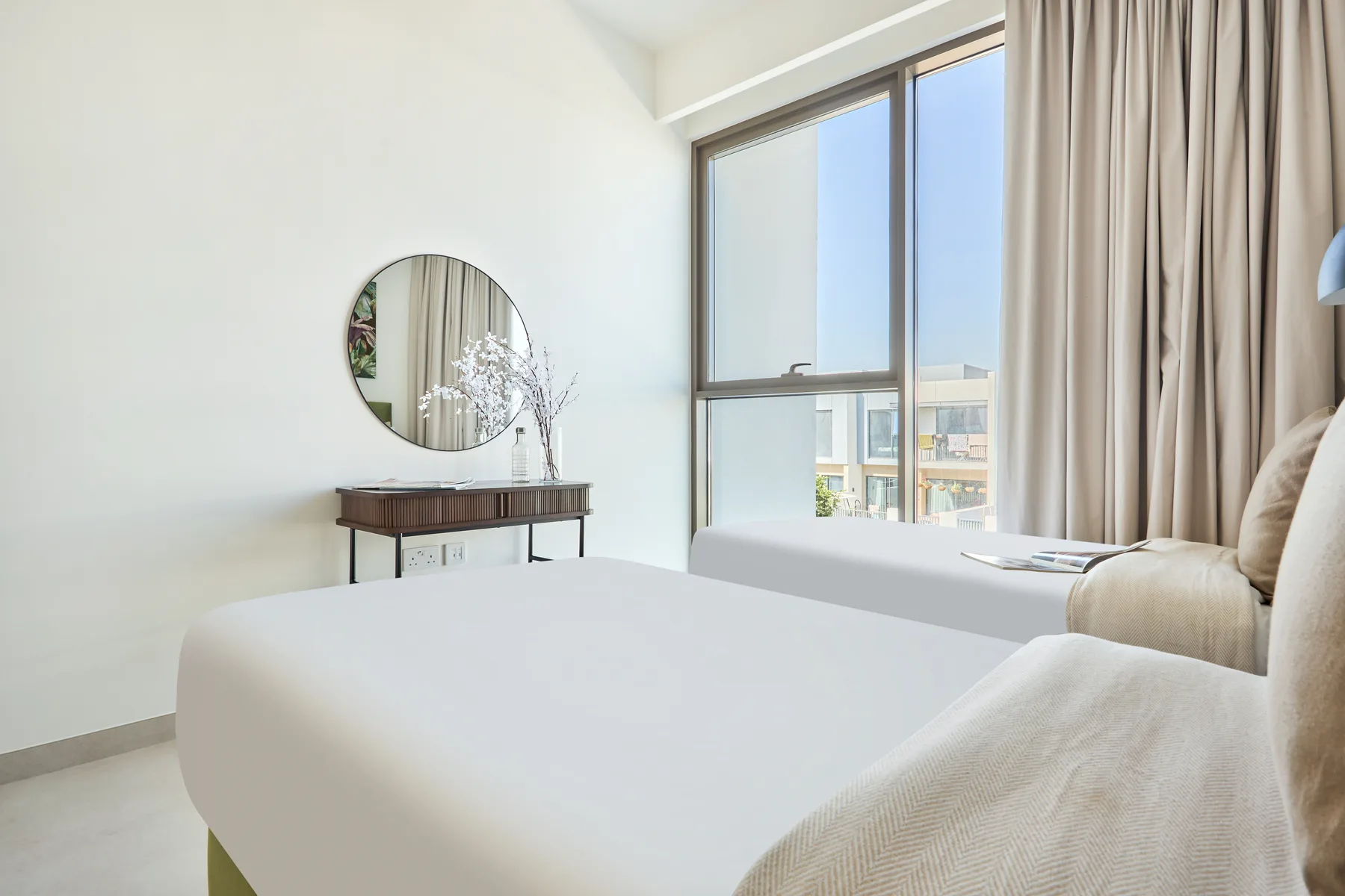 A bright bedroom with white bedding, a round mirror, and a large window.