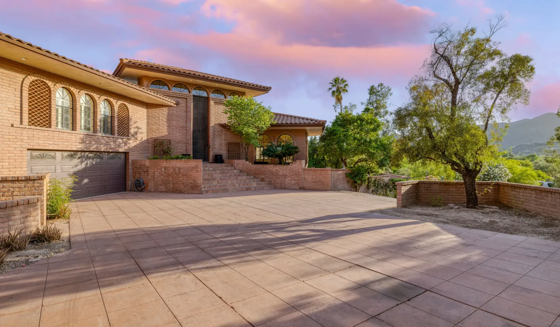 Una casa grande da a un patio pavimentado con árboles y colinas a lo lejos.