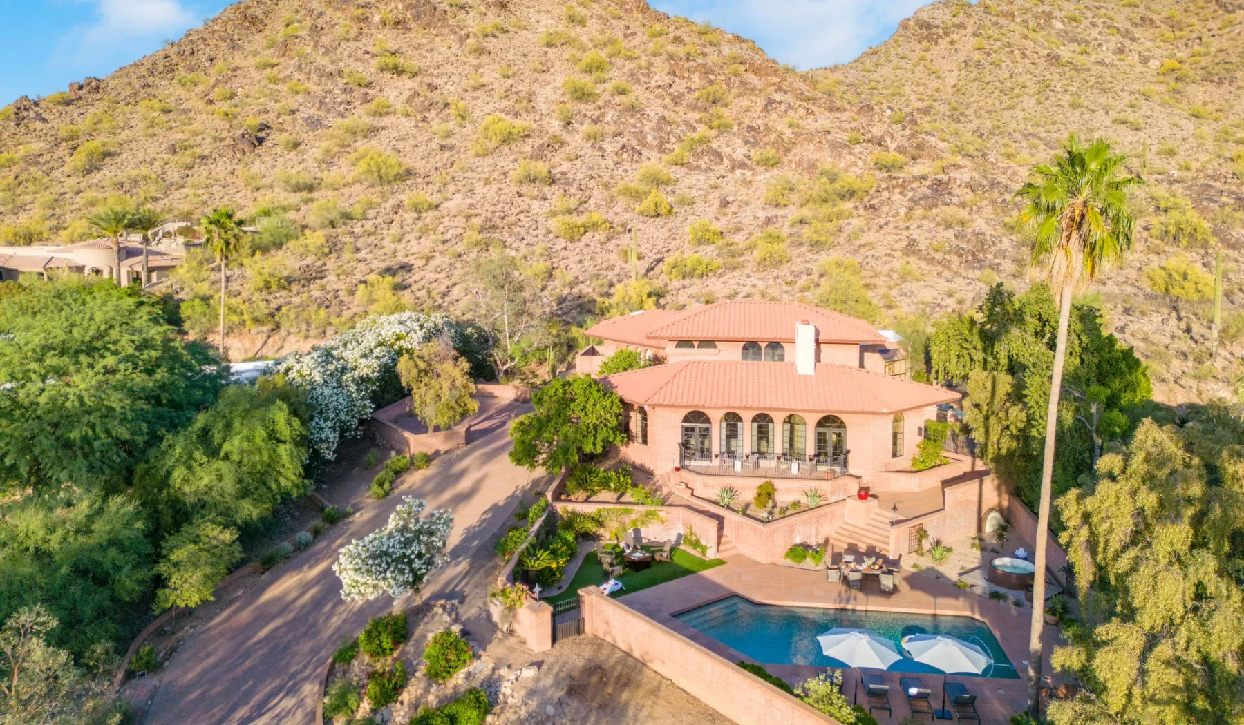 Una casa en la ladera domina una zona ajardinada con piscina bajo colinas rocosas y áridas.