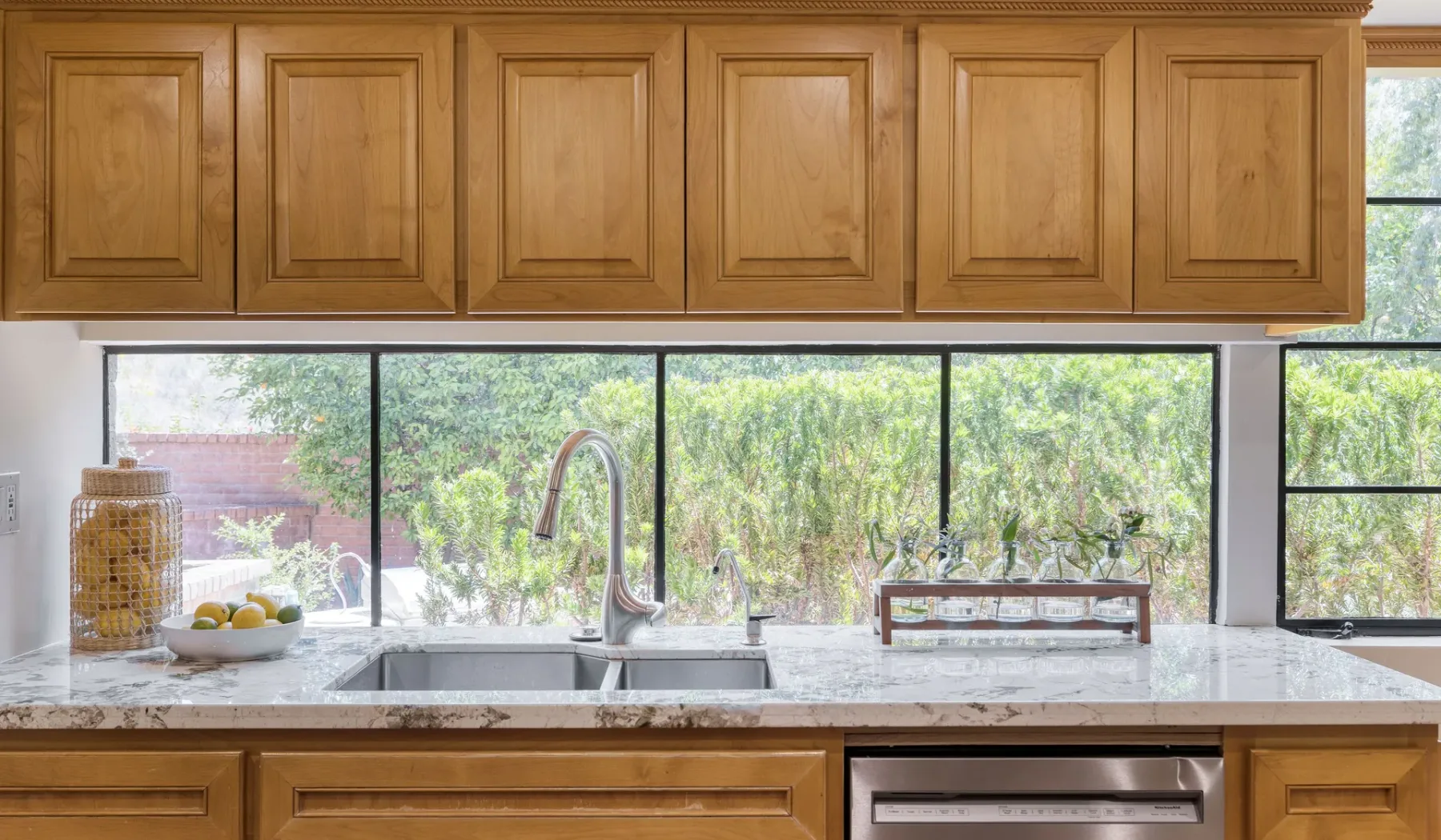 Una encimera de cocina con fregadero está bajo armarios de madera y amplias ventanas.