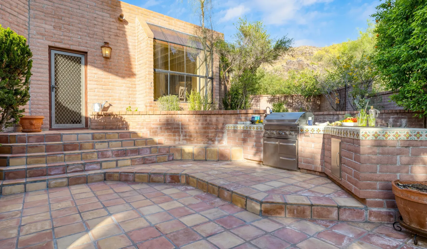 Un patio exterior embaldosado con una parrilla empotrada y vegetación alrededor.