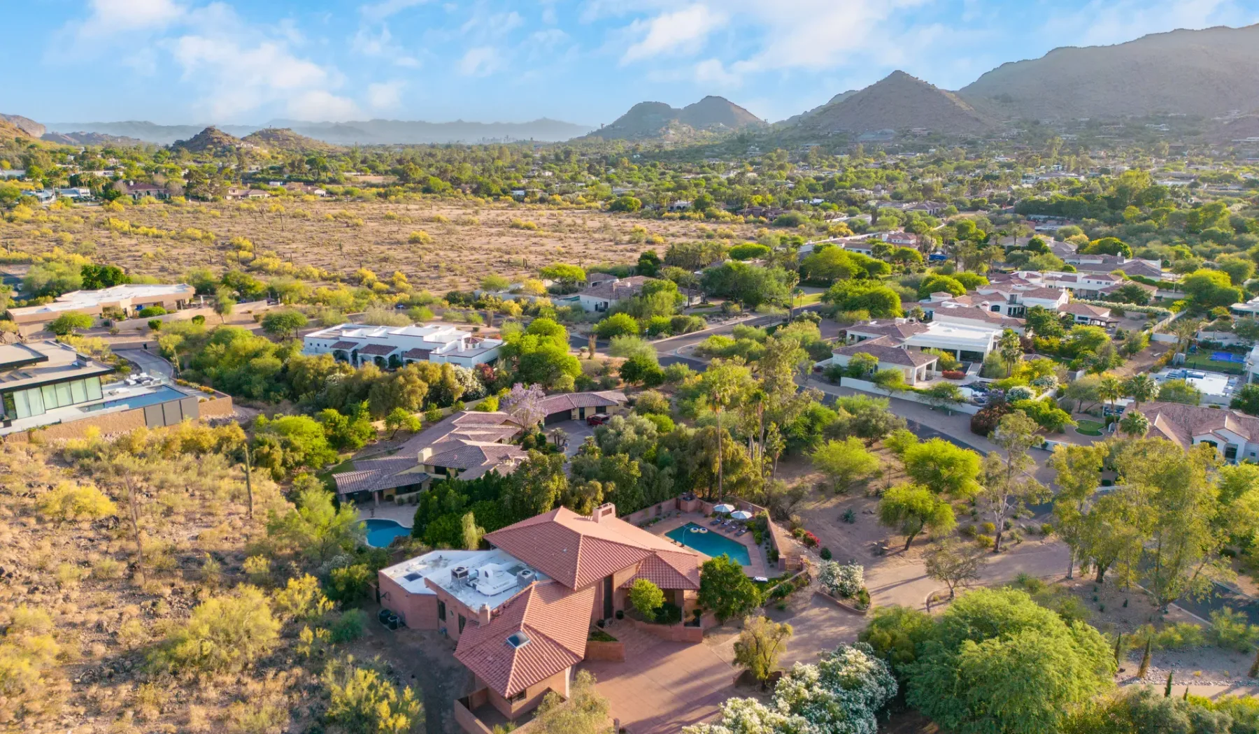 Vista aérea de una vivienda y edificios cercanos entre vegetación seca, con montañas al fondo.
