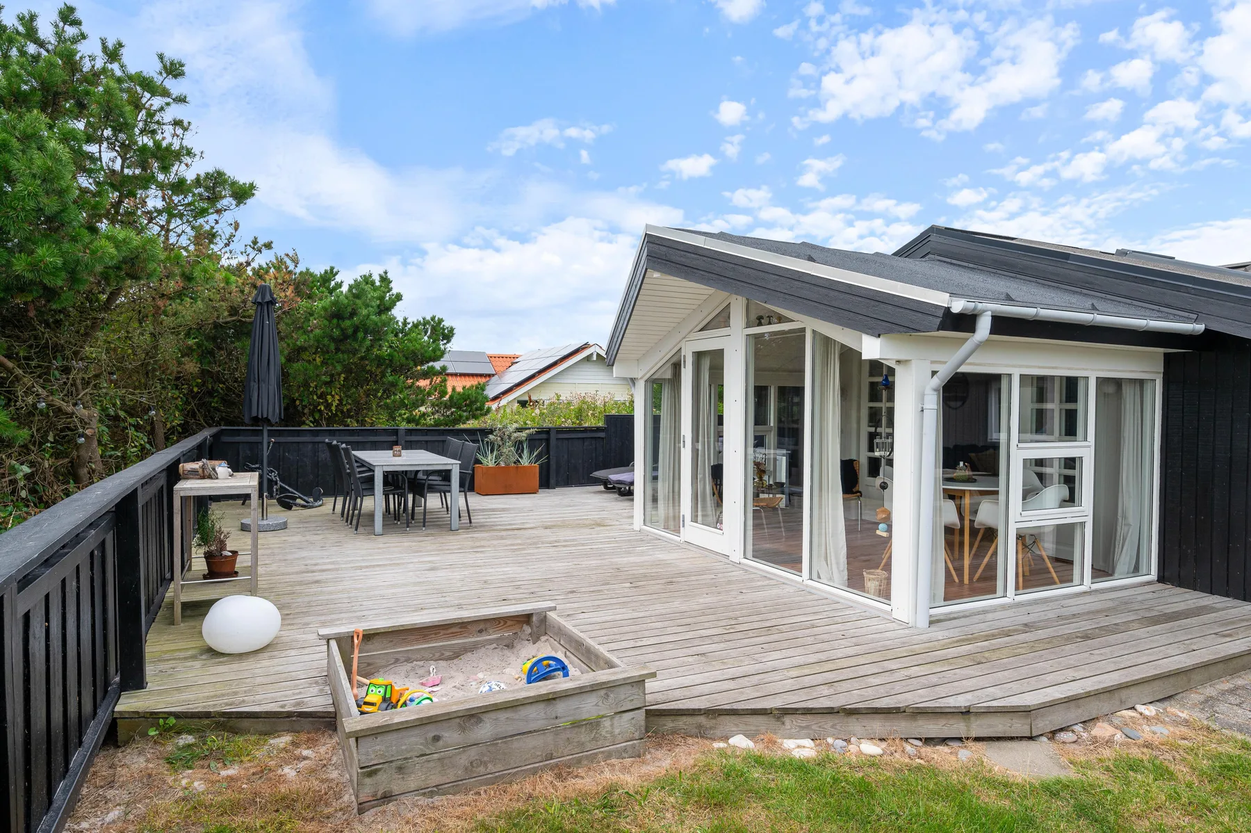 A house opens onto a wooden deck with outdoor seating and a small sandbox.