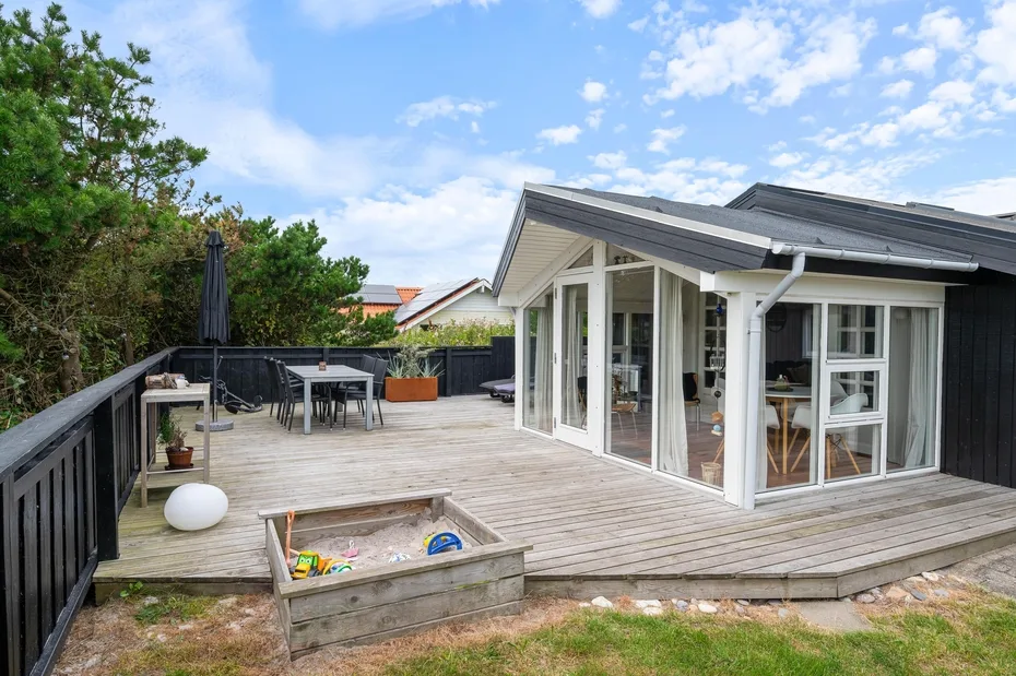 A raised wooden deck beside a glass-walled room, with outdoor seating and trees.