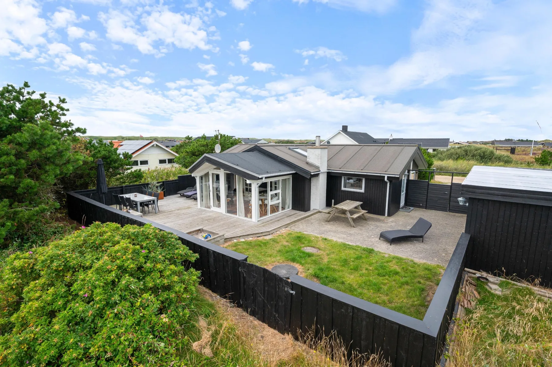 A modern house with a patio and fenced yard sits among low greenery.