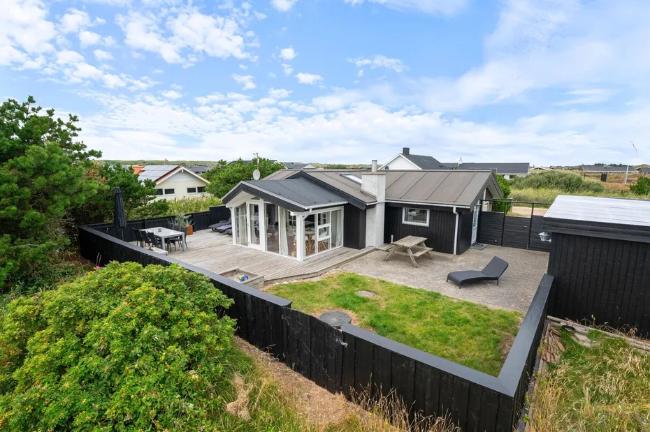 A low modern house sits in a fenced yard with a small patio and outdoor seating.