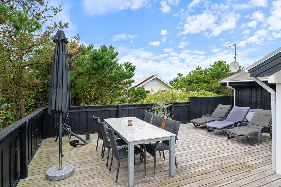 An outdoor deck has a dining table, seating, and lounge chairs beneath a blue sky.