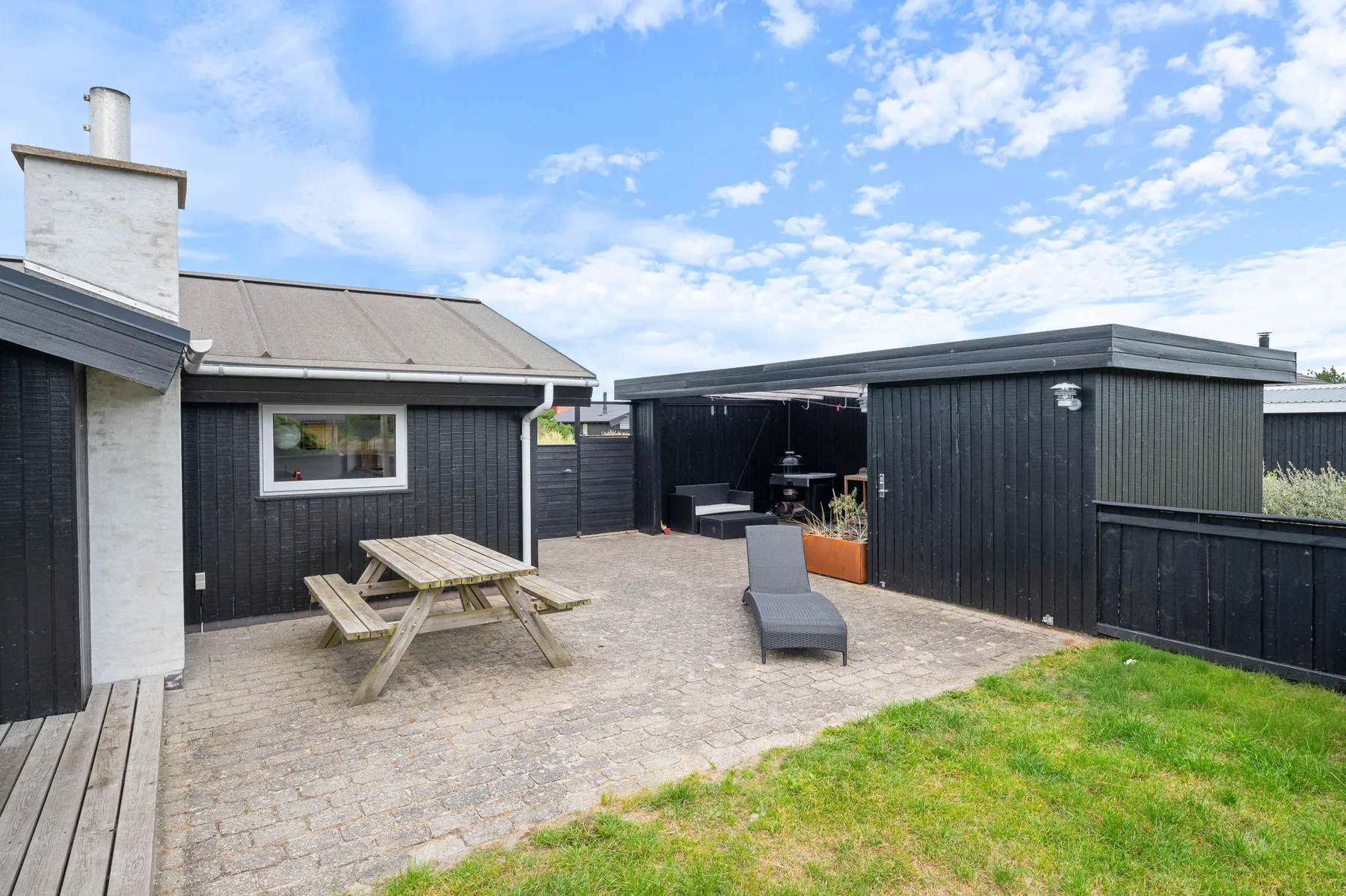 An outdoor patio with a picnic table and lounge chair beside dark-sided buildings.