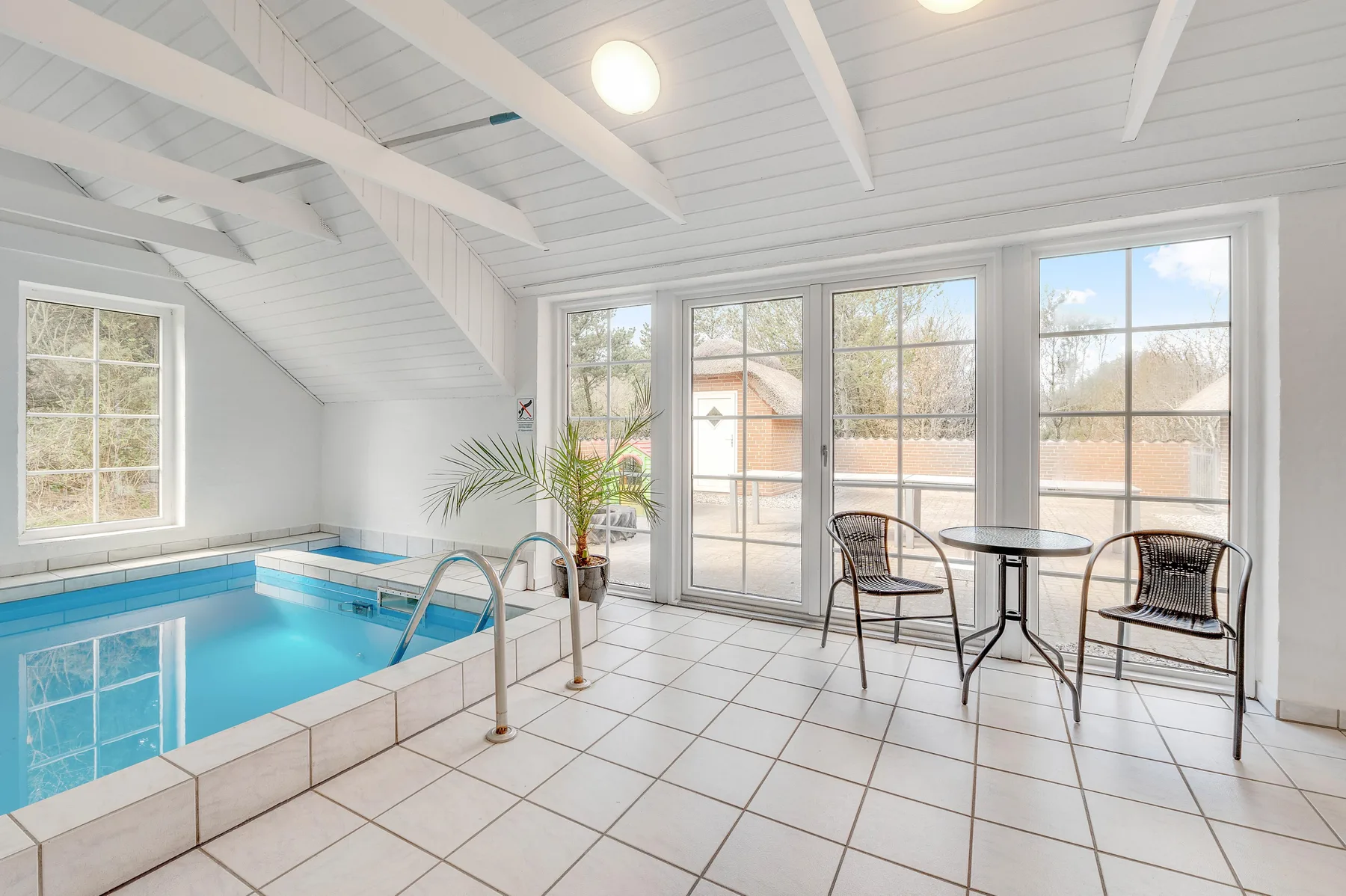 Una zona de piscina cubierta con suelo de baldosas y asientos junto a grandes ventanales.
