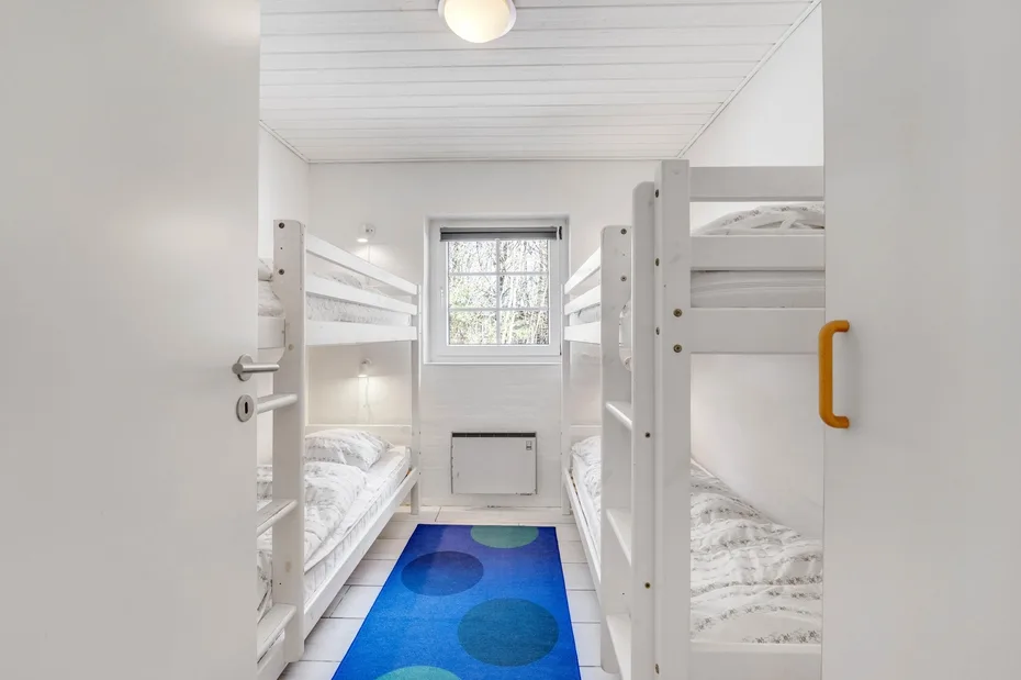 Un dormitorio luminoso con literas blancas y una alfombra azul bajo la ventana.