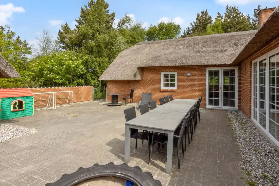Un patio pavimentado junto a una casa de ladrillo, con una larga mesa de comedor al aire libre.