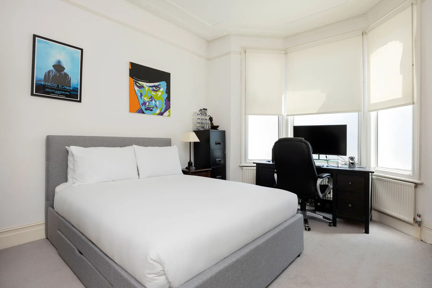 A bedroom with a neatly made bed and a desk beside the windows.