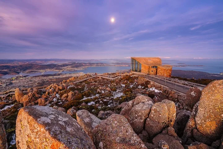 A rocky lookout overlooks a broad bay and a small building beneath a colorful sky.