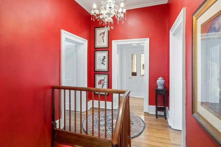 A red-walled hallway with a wooden railing, framed artwork, and a chandelier.