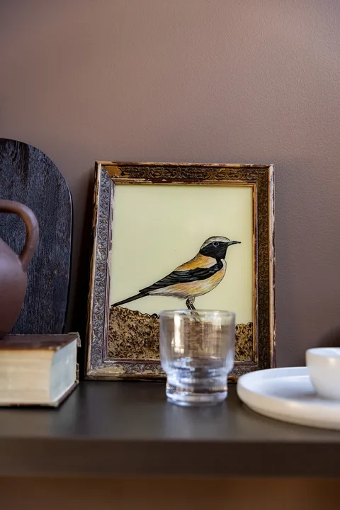 A framed bird illustration sits on a shelf beside a glass and dishes.