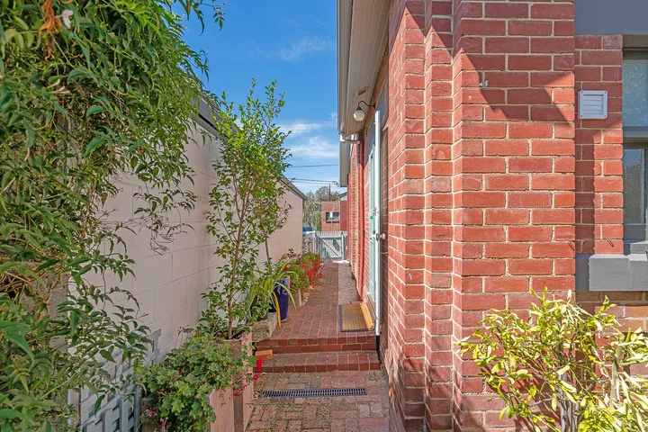 A narrow brick walkway runs beside a red brick building, with plants along the path.