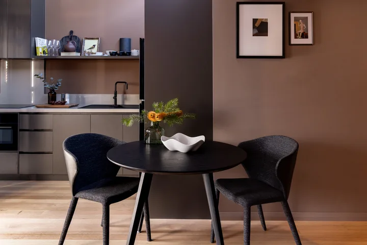 A dining area with a round table and dark upholstered seating beside a compact kitchen.