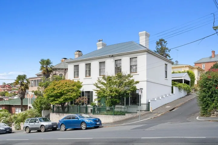 A white two-story building sits on a sloping street among trees and parked cars.