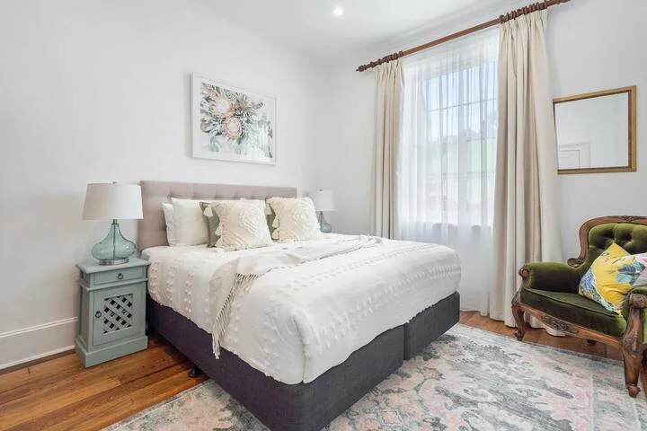 A bright bedroom with a neatly made bed beside a curtained window.