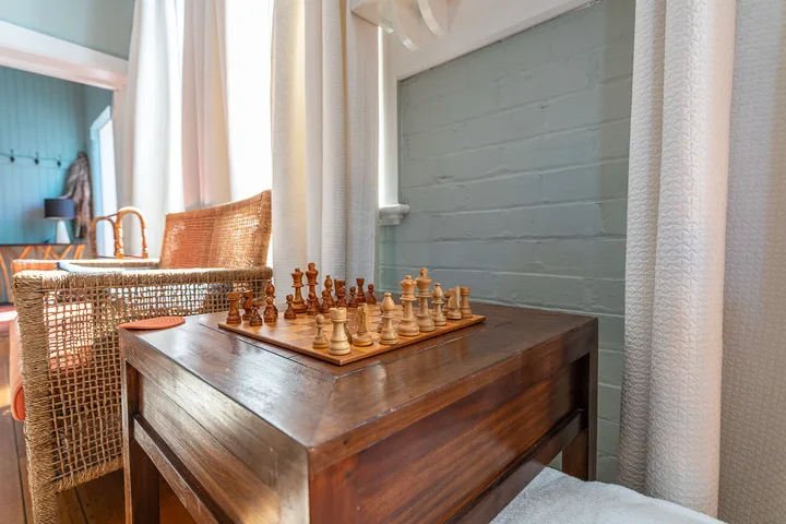 A wooden table with a chessboard sits beside wicker seating and pale curtains.