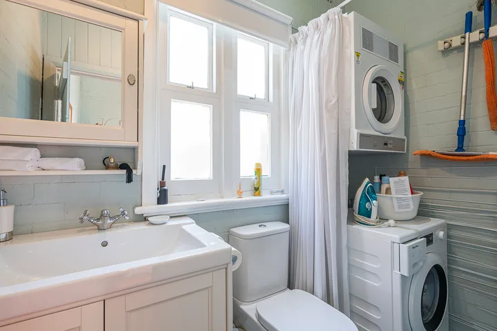 A bright bathroom with a sink, toilet, and stacked laundry appliances beside a window.