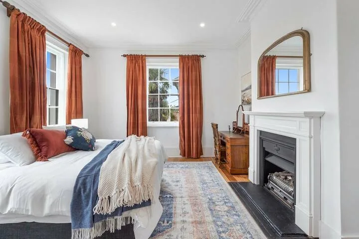A bright bedroom with a bed, rust-colored curtains, and a fireplace.