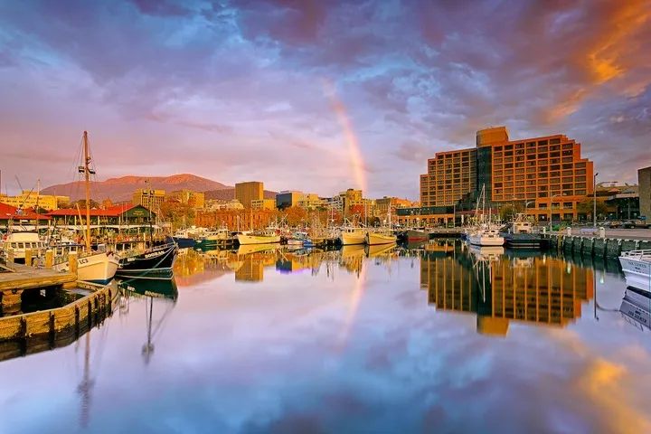 A marina with moored boats reflects the waterfront buildings and colorful evening sky.