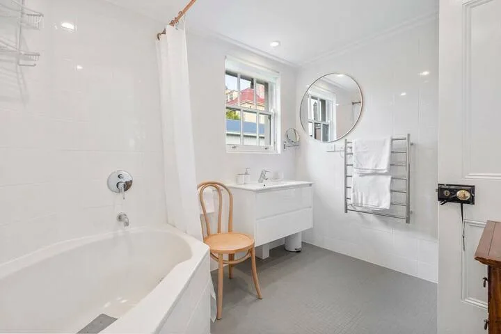A bright white bathroom with a bathtub, vanity, and round wall mirror.