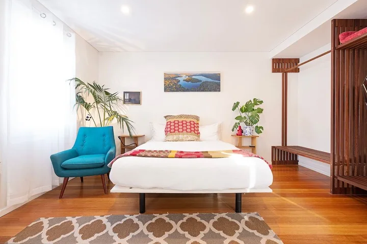 A bright bedroom with a white bed, turquoise seating, and wooden flooring.