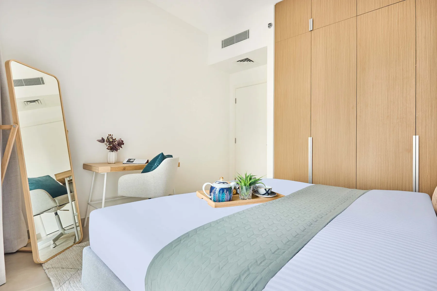 A bright bedroom with a neatly made bed and a small desk beside a mirror.