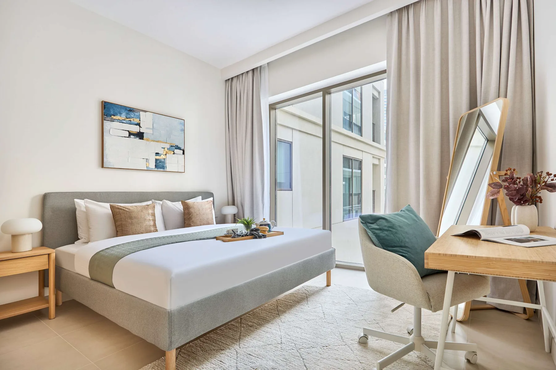 A bright bedroom with a neatly made bed beside large windows and light curtains.