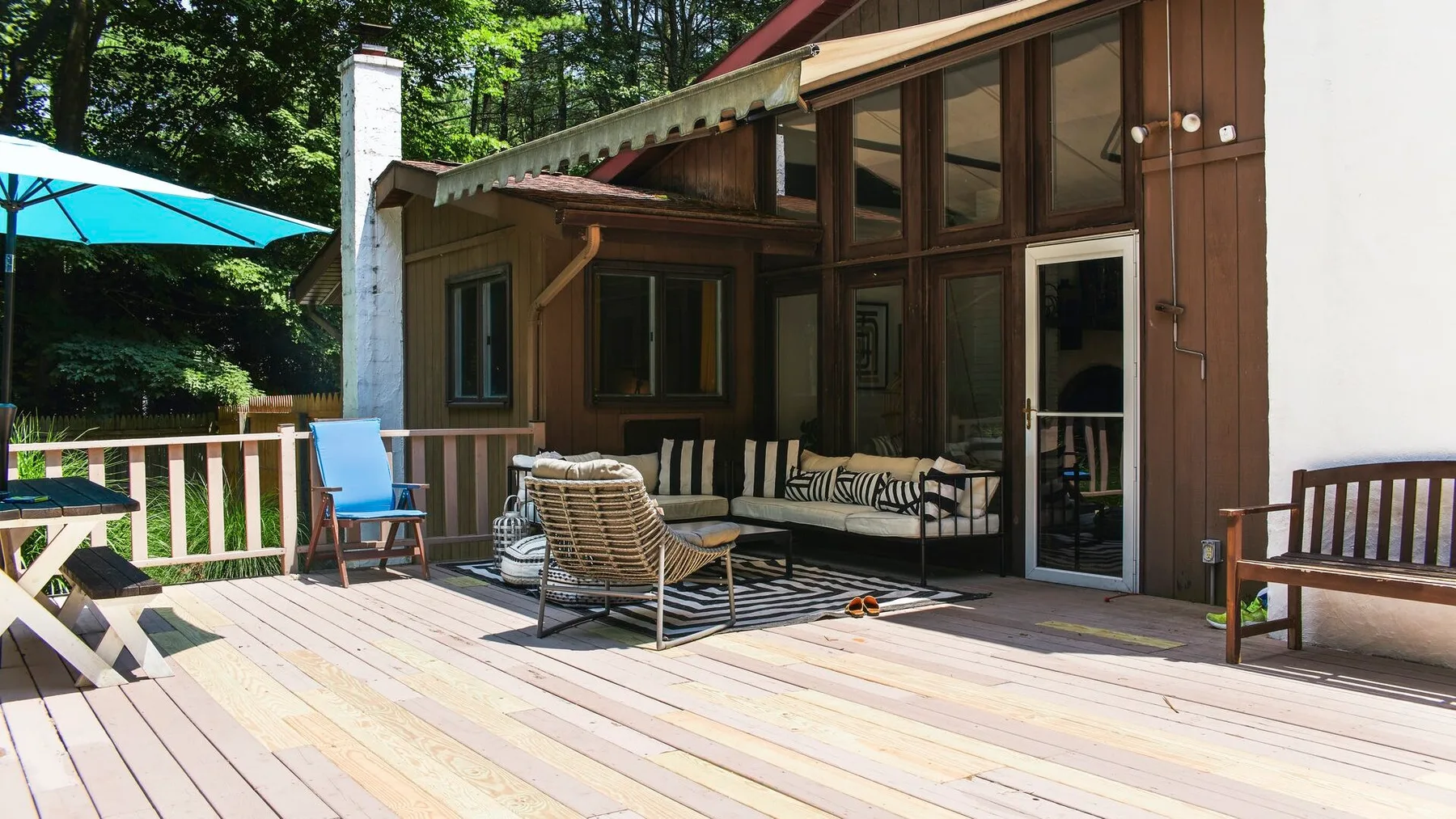 Una terraza de madera junto a una casa tiene asientos al aire libre y una sombrilla turquesa.
