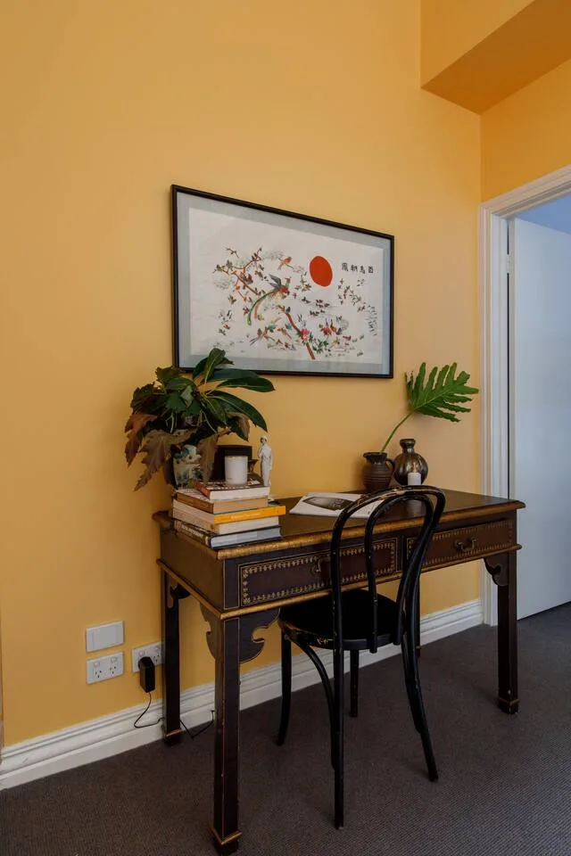 A writing desk with a chair and framed artwork sits against a yellow wall.