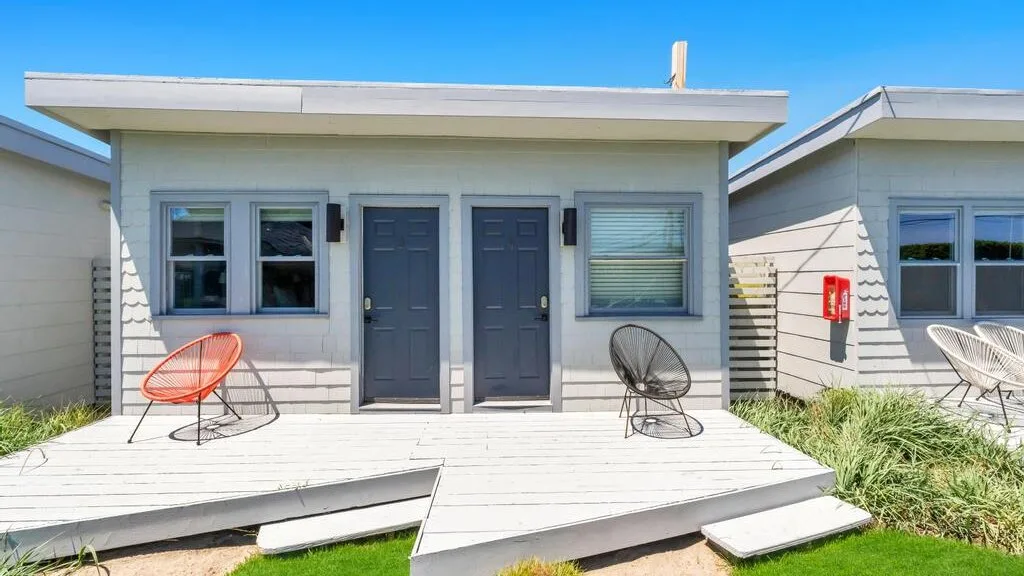 A small building opens onto a white deck with two dark doors and outdoor seating.