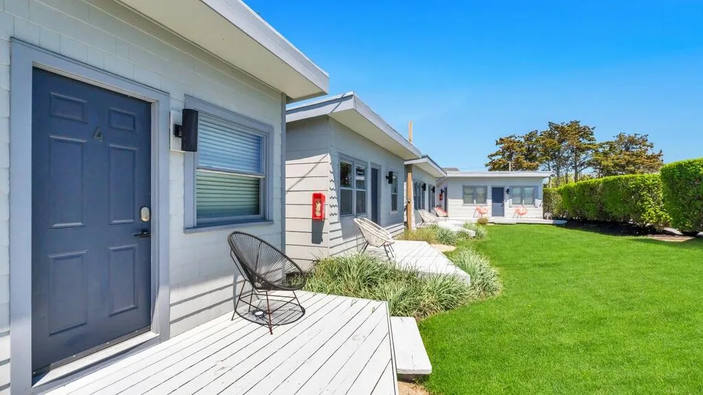 A row of small buildings opens onto a green lawn, with a chair on the deck.