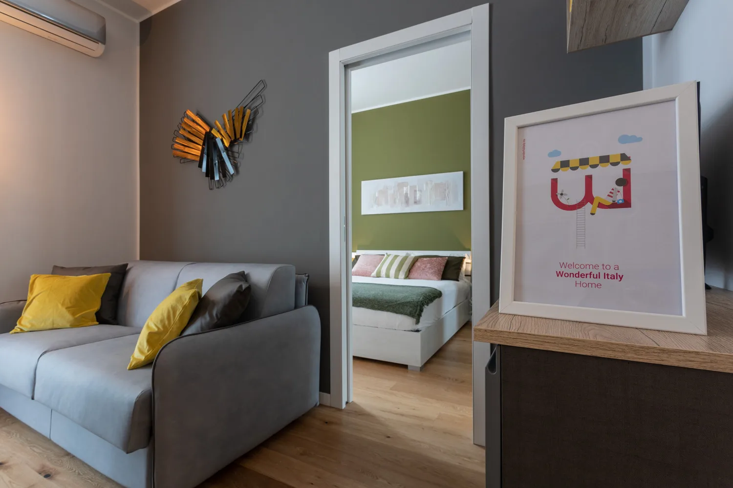 Una sala de estar con asientos grises y una vista de un dormitorio a través de una puerta abierta.