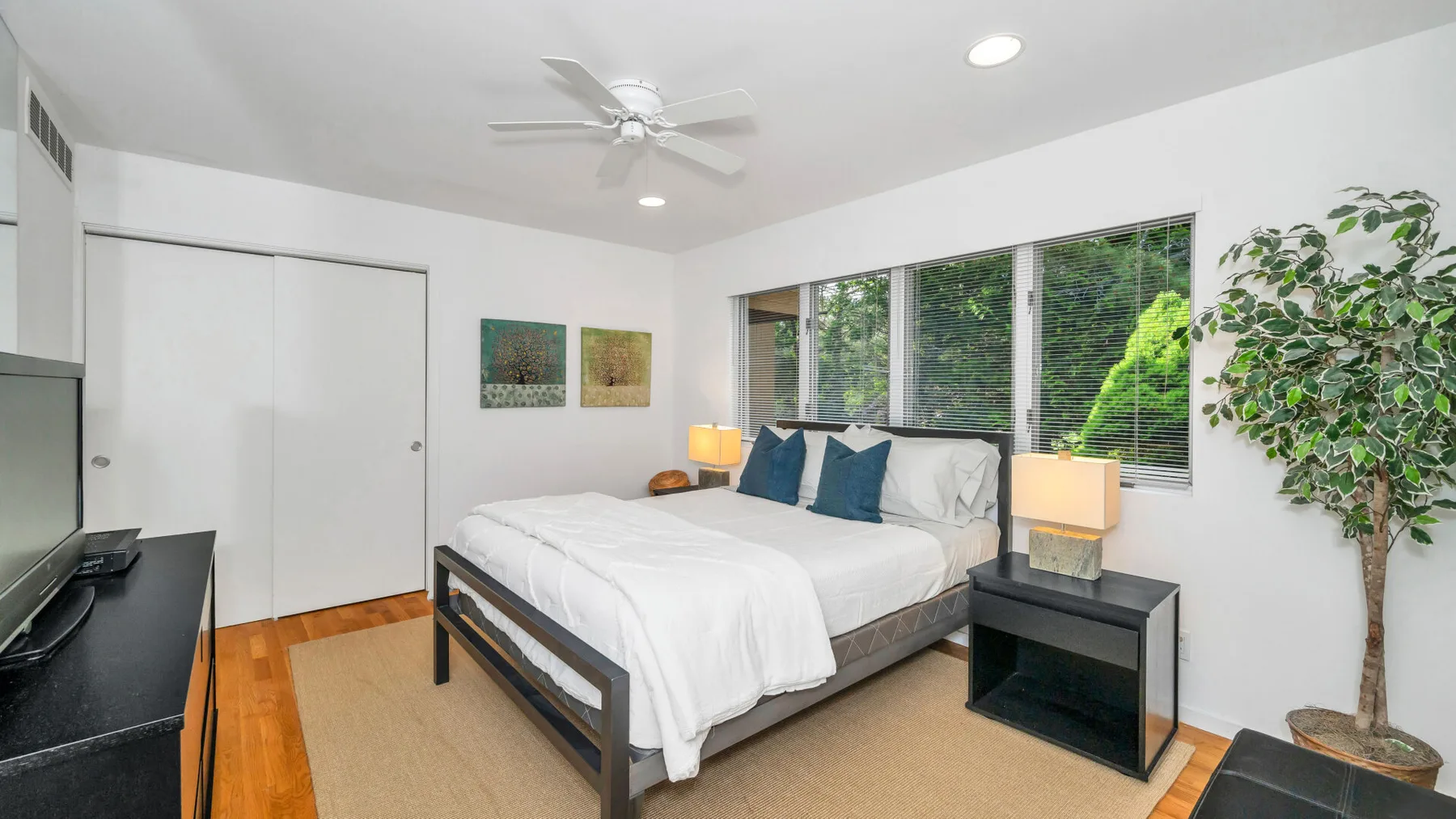 Un dormitorio luminoso con una cama junto a grandes ventanas y una planta frondosa.