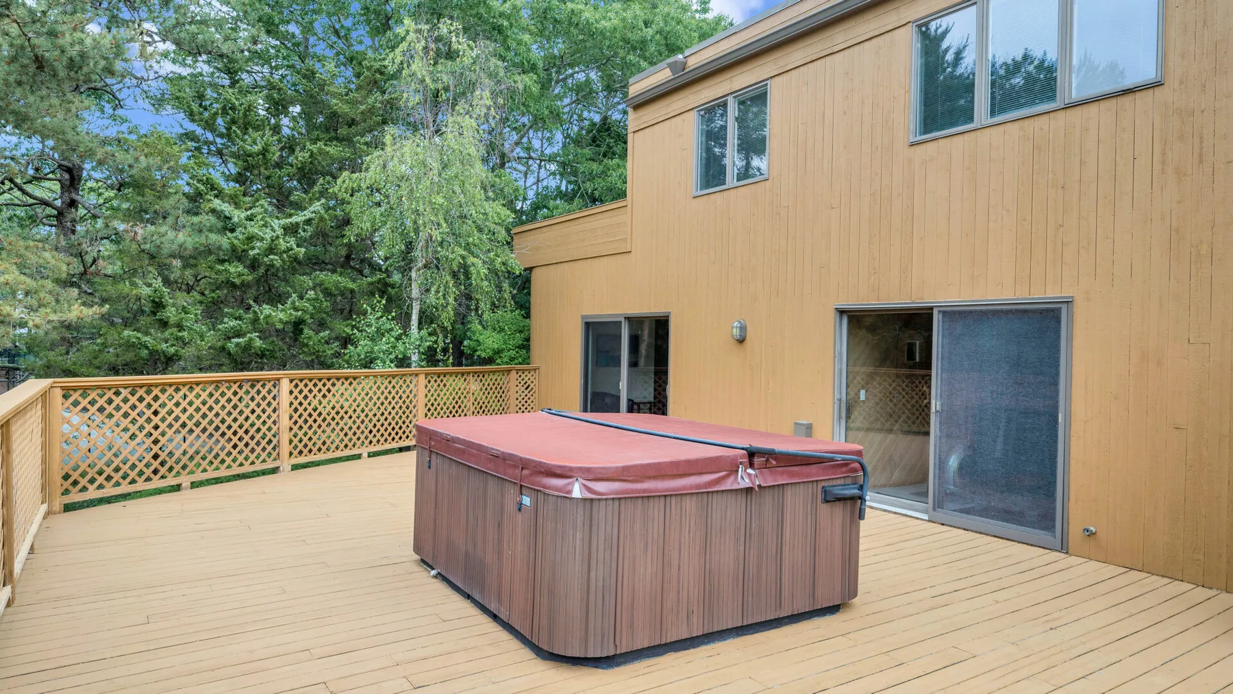Una terraza de madera junto a un edificio color beige tiene una bañera de hidromasaje y una barandilla de celosía.