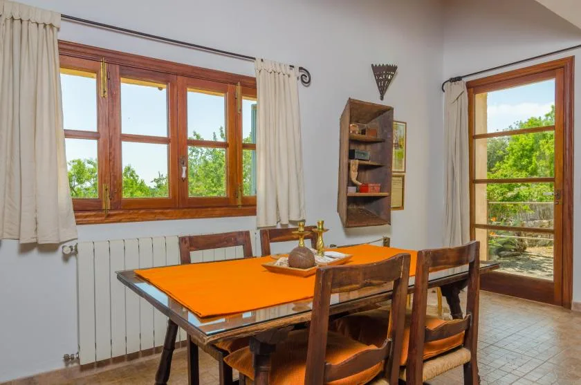 Comedor con una mesa de madera bajo grandes ventanas y junto a una puerta abierta.
