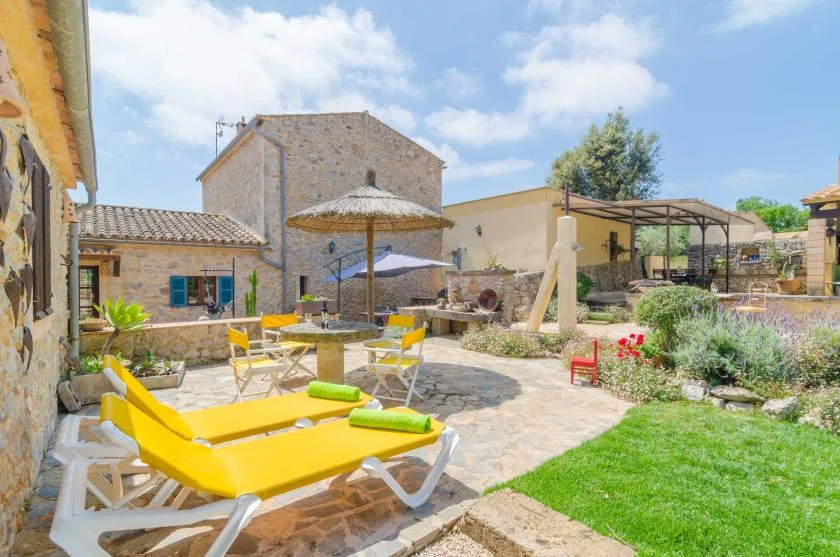 Un patio soleado de piedra con tumbonas amarillas, una sombrilla y una zona de césped.