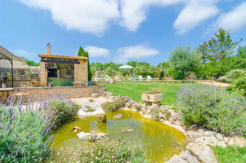Un estanque de jardín está rodeado de plantas con flores, con asientos al aire libre y un pequeño bar cerca.
