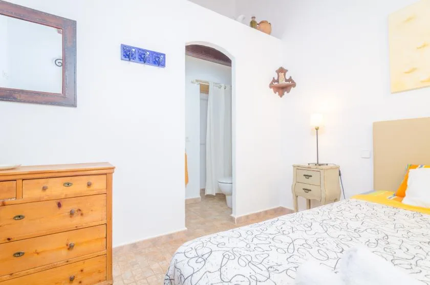 Un dormitorio con una colcha estampada, una cómoda de madera y una puerta hacia un baño.