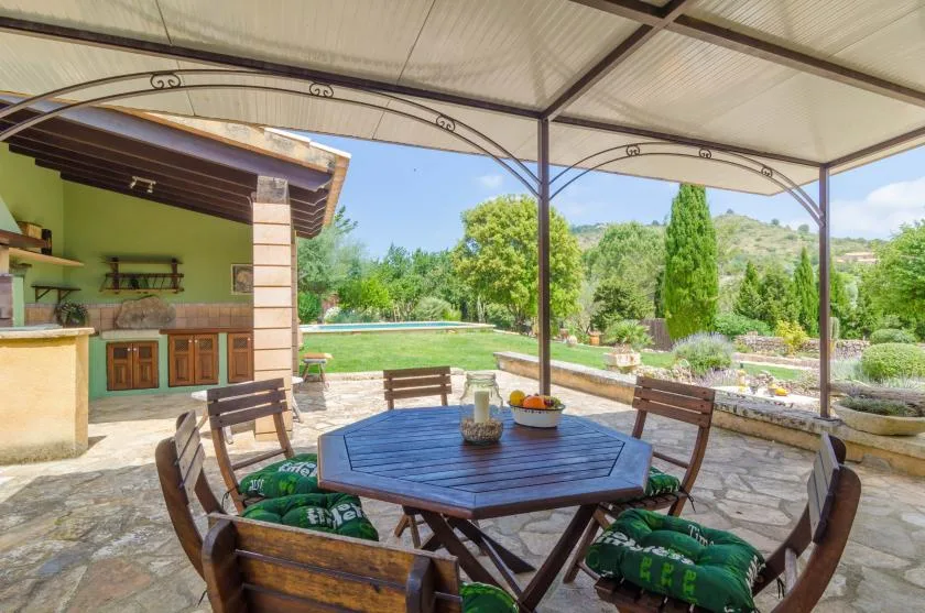 Un patio exterior cubierto tiene una mesa de madera y asientos frente a un jardín.