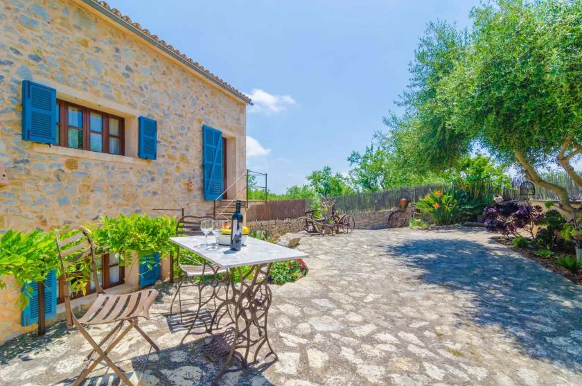 Una casa de piedra da a un patio pavimentado con asientos al aire libre y árboles frondosos.