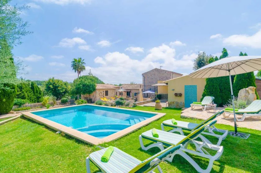 Una piscina está junto a un jardín soleado con tumbonas y una sombrilla.