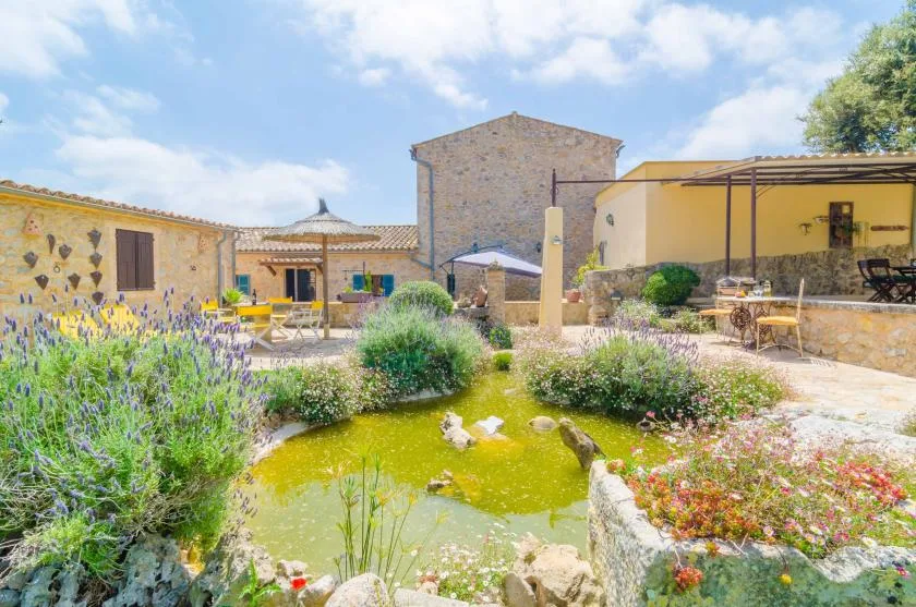 Un jardín soleado rodea un pequeño estanque, con edificios de piedra al fondo.