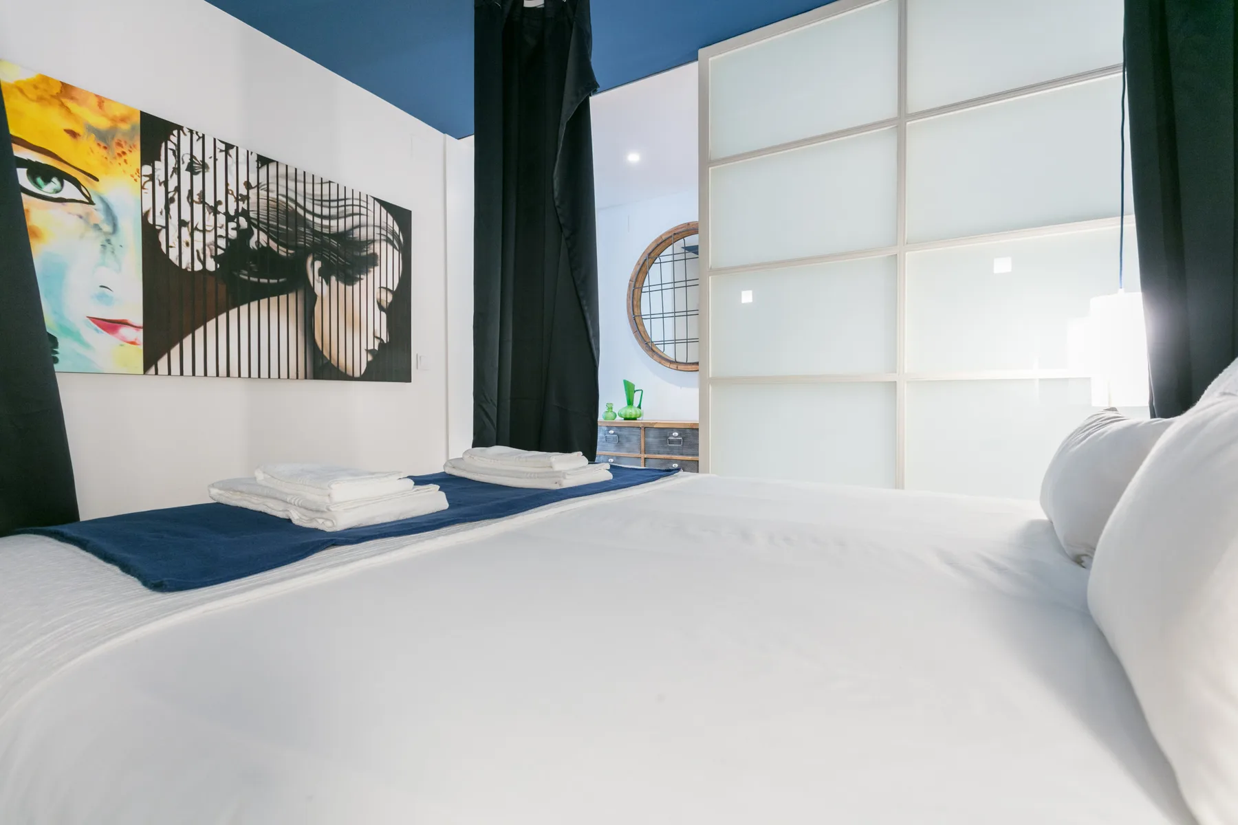 Un dormitorio con ropa de cama blanca, arte colorido en la pared y un separador de vidrio esmerilado.