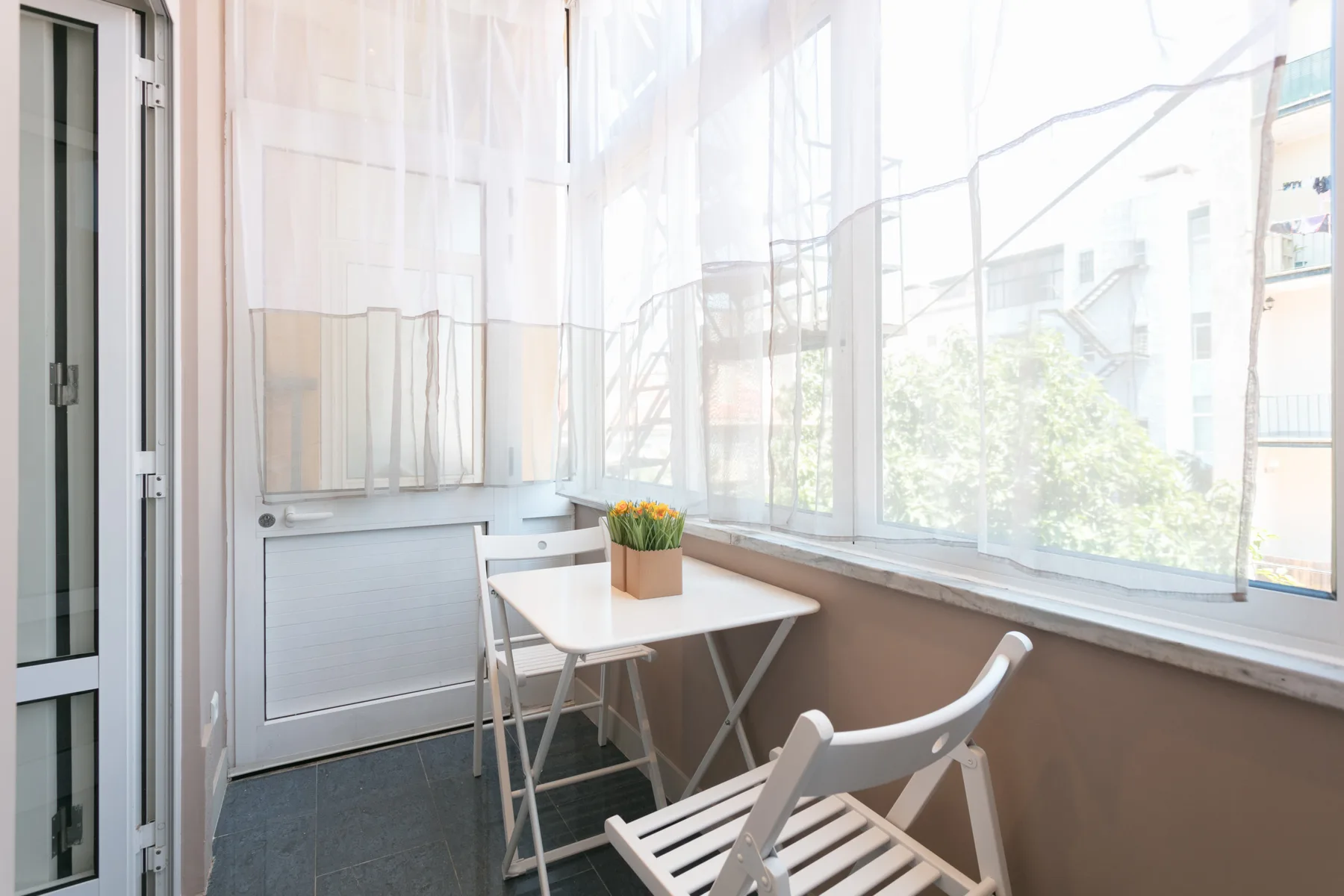 Un balcón cerrado y luminoso con una mesa pequeña y asientos junto a grandes ventanas.